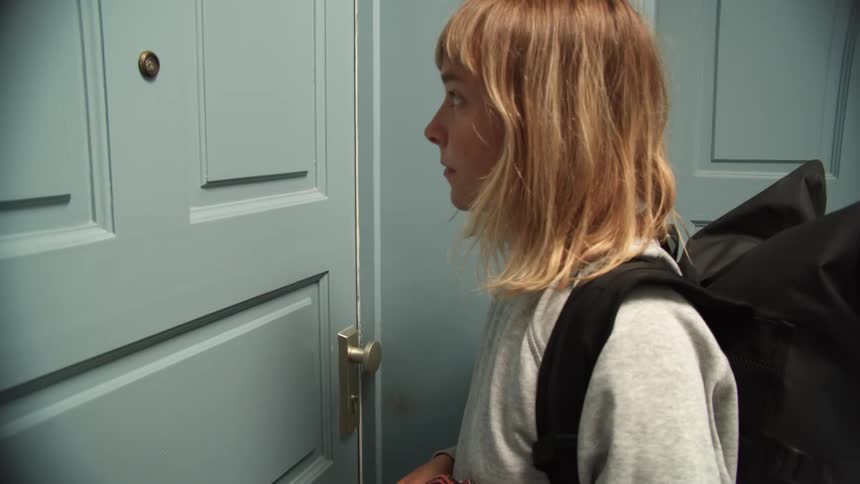 A young woman wearing a black backpack walks up to blue apartment door and knocks on it lightly