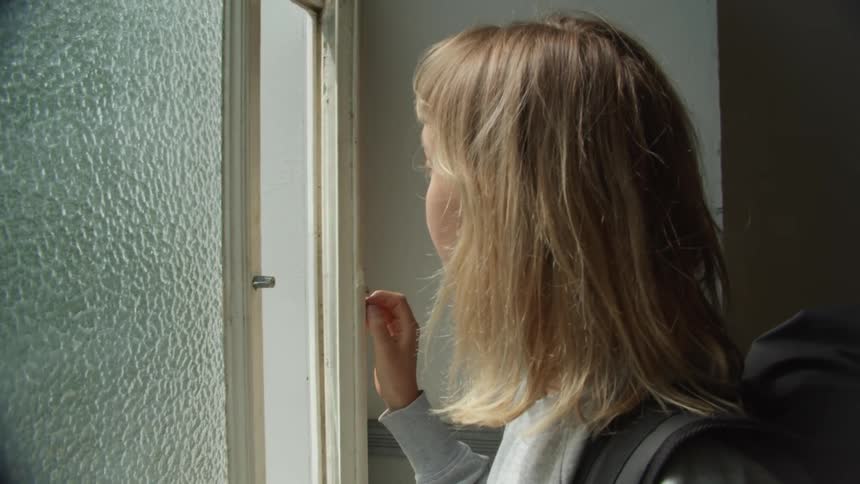 A woman in a room with a sweater and a backpack opening a window