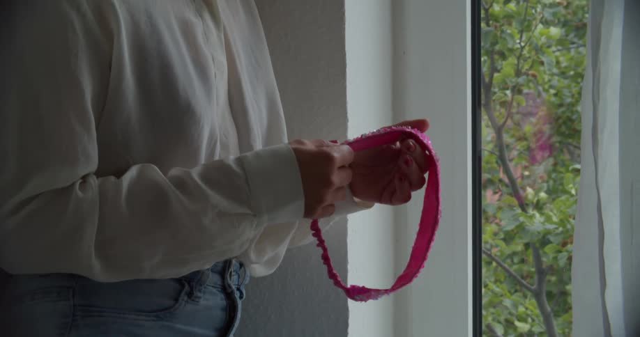 A woman standing indoors looking out a window rubs her fingers over a pattern on a pink headband at daytime