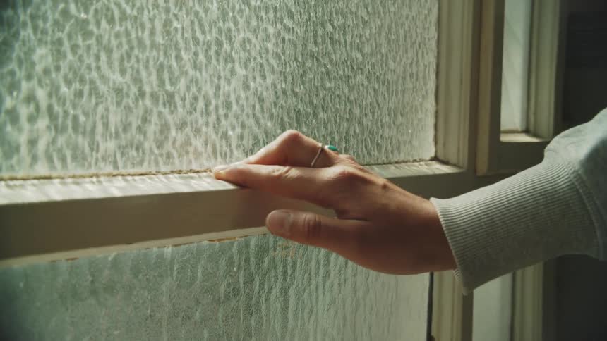 A female hand with rings on the fingers sliding over the frame of a window pane inside a room