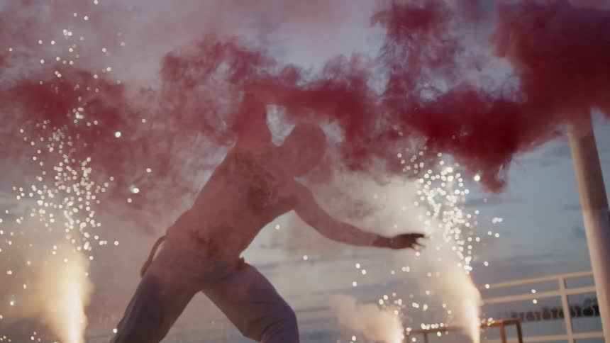 A man is outside doing a cartwheel and capoeira-dancing among fireworks and red smoke
