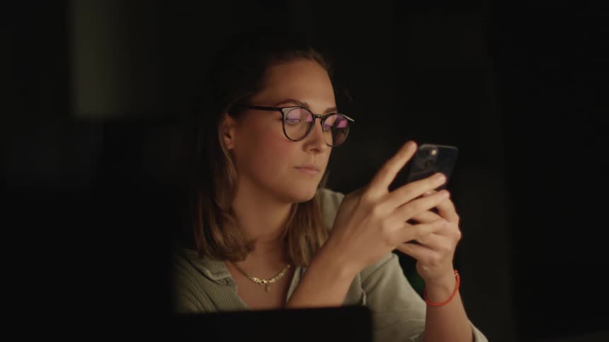A woman is sitting in a nearly dark room and looking on her cell phone