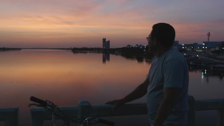 Man with bicycle stands near rail looking over river reflecting red sky with town in in the distance