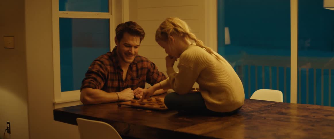 A young girl is sitting on the table as her father sits in a chair and they play a game of checkers