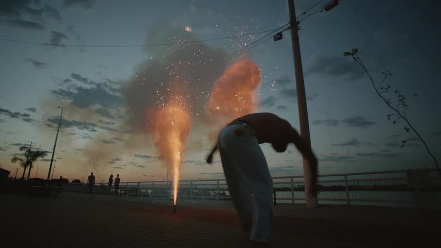 A silhouetted man kicking his leg while doing capoeira with fireworks exploding in the background