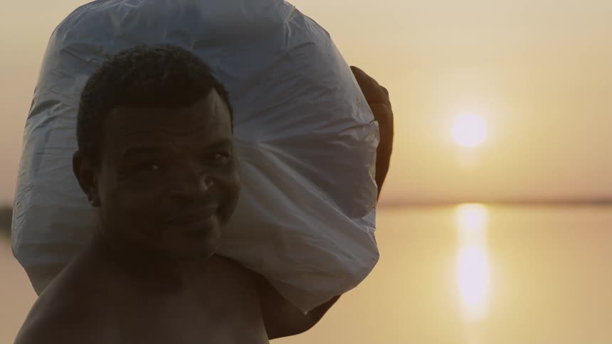 A man from Northeast Brazil with a large bag on his shoulder looks at the sun setting over the ocean and turns and smiles
