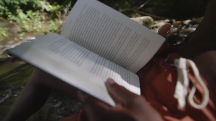 A boy reads a book while sitting along the river