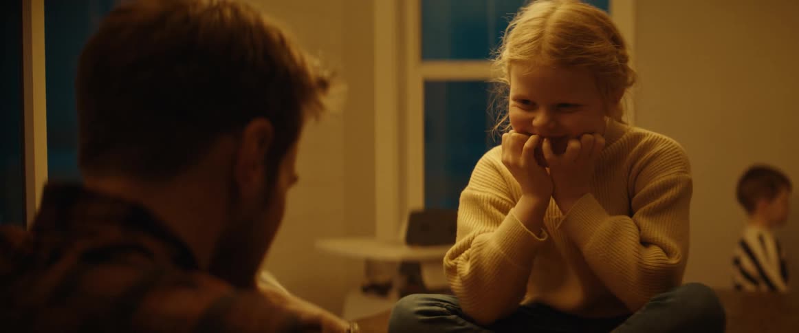 A young girl is sitting on the table as her father sits in a chair as they play a game of checkers and a little boy walks past them