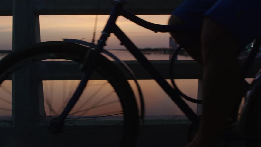 Man rides bicycle across the bridge at dusk with orange reflections in the river in the background