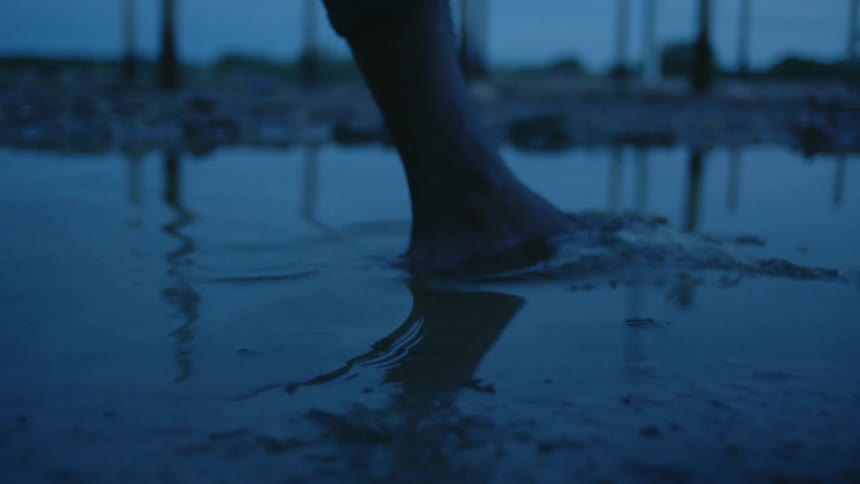 Close up of a barefoot person walking on a puddle of water outdoors at evening