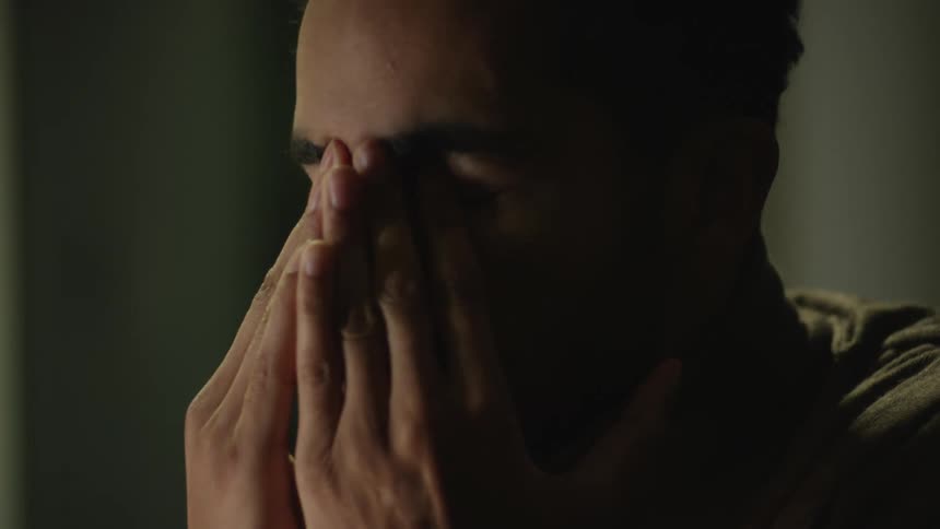 A man is sitting at a table and he rubs his face with his hands and closes his eyes as he folds his hands
