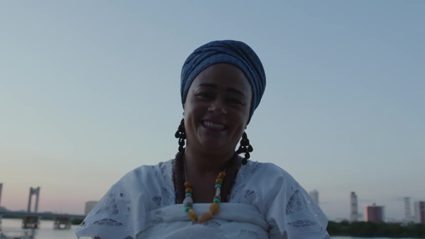 A woman from Bahia, Brazil, in a white dress and a turban is standing near water smiling and there are tall city buildings in the distance