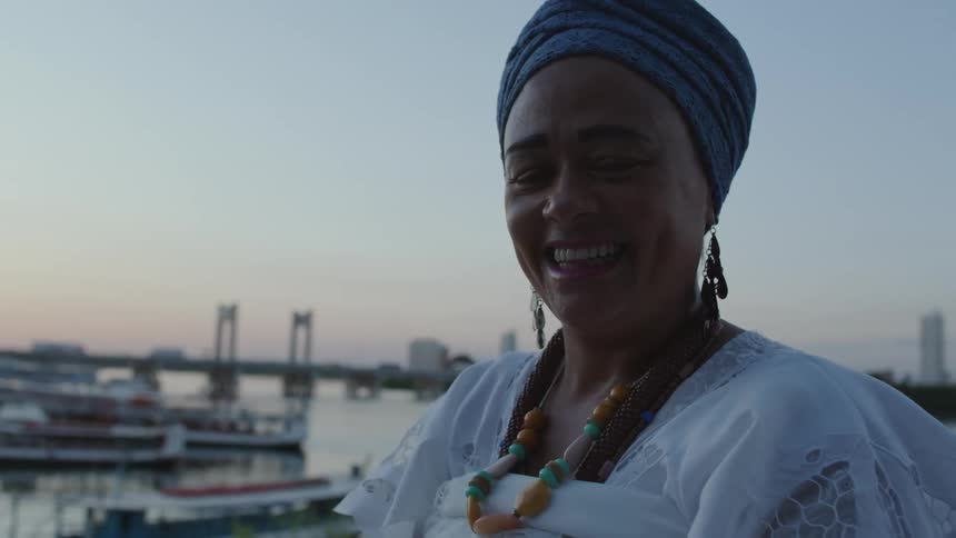 A woman in typical Baiana clothing stands on the waterfront with boats docked and smiles for a portrait