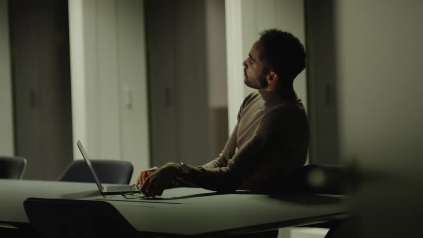 Man wearing brown sweater sits at desk working on laptop in room