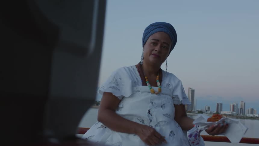 Woman wearing turban serves food while watching a television with water and the city skyline in the background