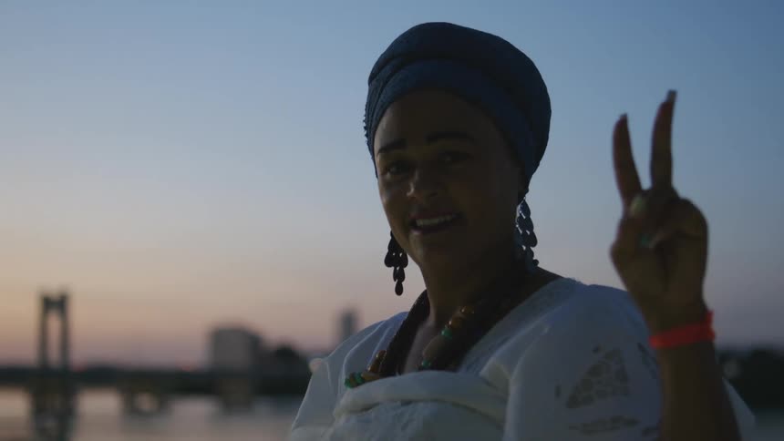 A woman from Bahia poses while holding up a peace sign in front of a river at dusk