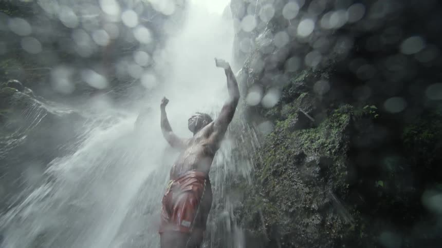 A man raises his arms and laughs while standing under a waterfall