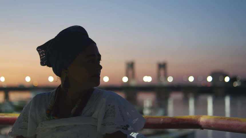 A woman is standing along the shore of a river with traffic moving as the bridge lights up in the background
