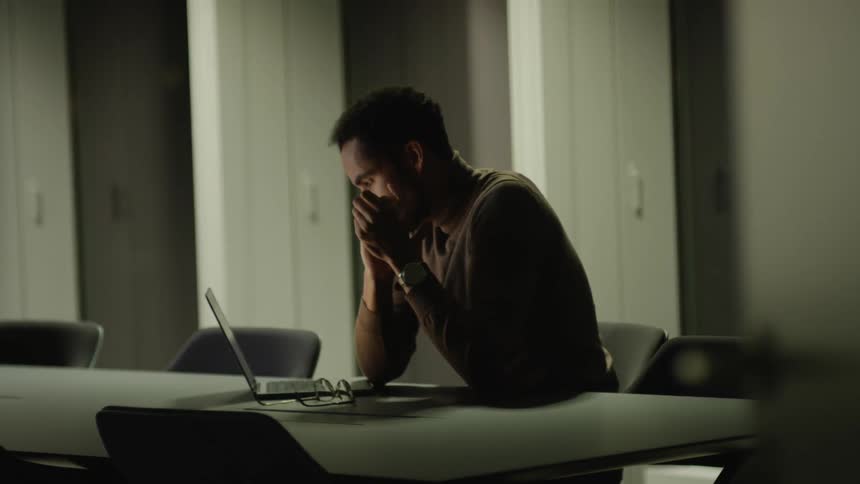 A man is sitting at a table with a laptop computer and his hands on his face as he puts on his glasses and types on the keyboard
