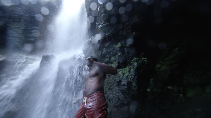 A man in swim trunks runs his hands through his hair while standing under a small waterfall during the day