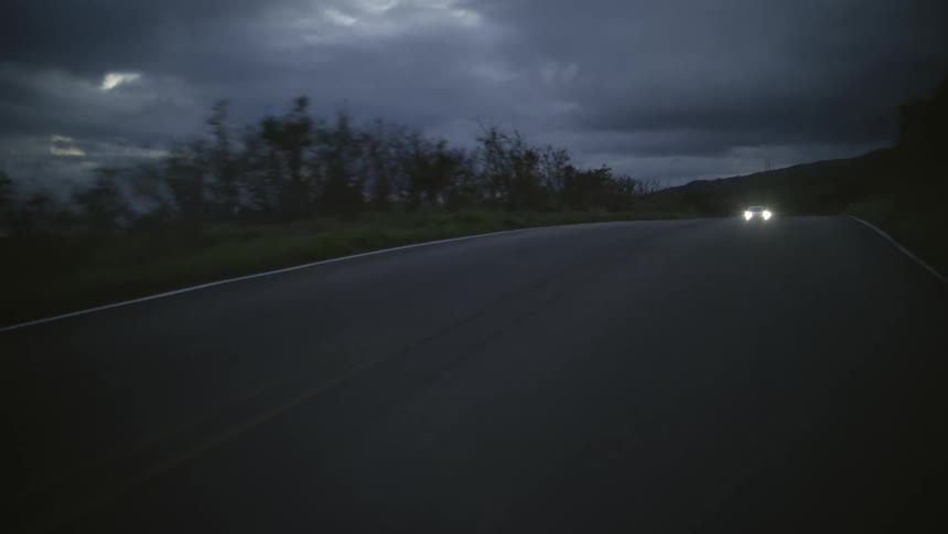 A white vehicle with headlights on drives on a road through the countryside at evening