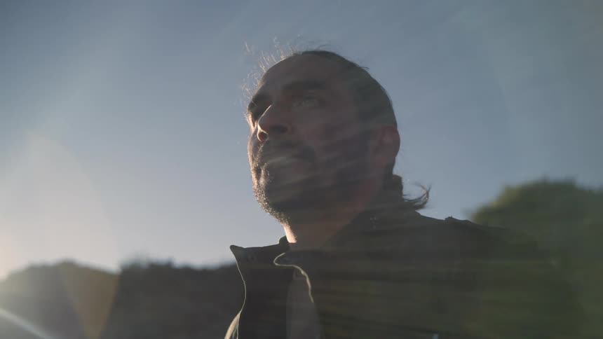 Man with beard long hair and black coat looks around near a hill