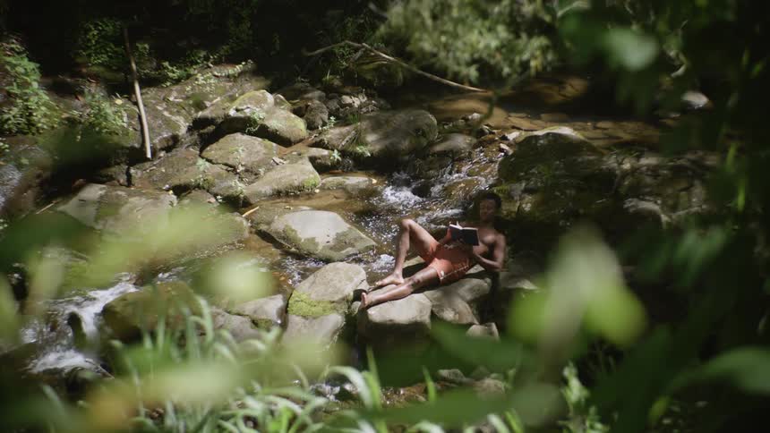 A man is lying on the rocks reading a book next to a flowing stream