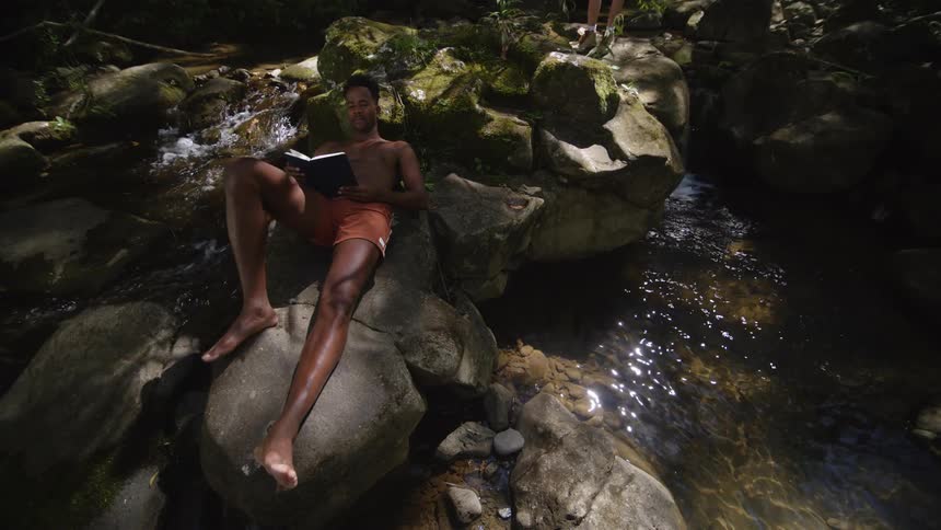A man in swim trunks reads a book while resting on top of a rock formation in a stream during the day