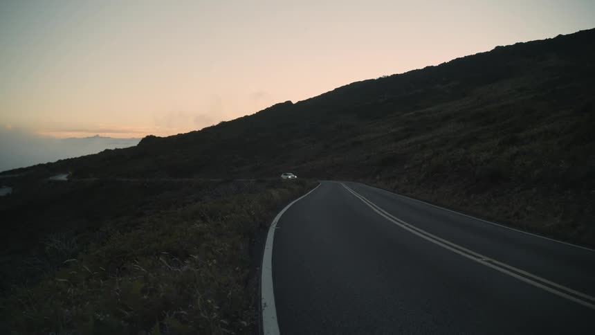 A white vehicle with headlights on drives on a road through the countryside at evening