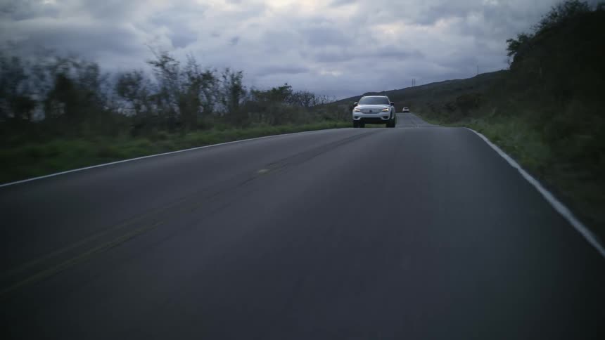 Automobiles travel on a road surrounded by plants and trees under a cloudy sky