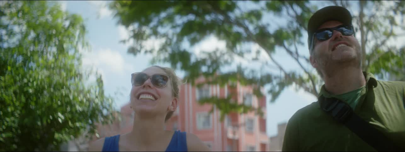 Couple stands smiling in park near pale colored apartment buildings