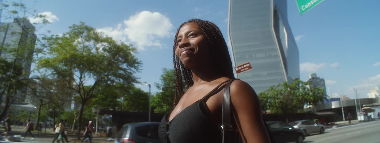 A woman smiles at what she's looking at while standing on the side of a downtown street as cars pass by and tall buildings are in the distance