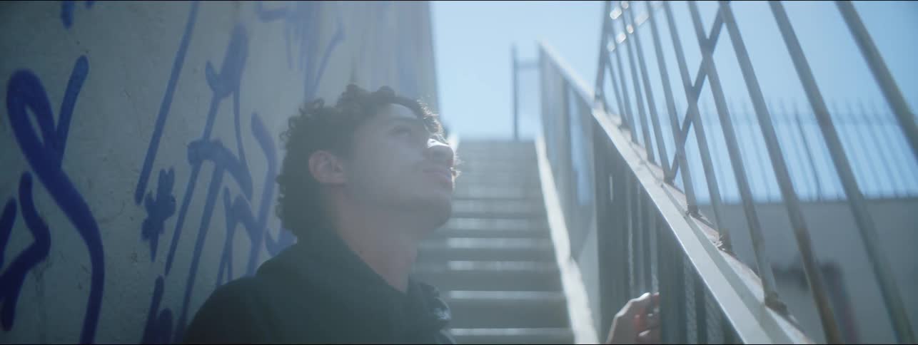 The sun shines brightly as a teenage boy stands on a staircase outside next to a graffiti painted wall as he looks up