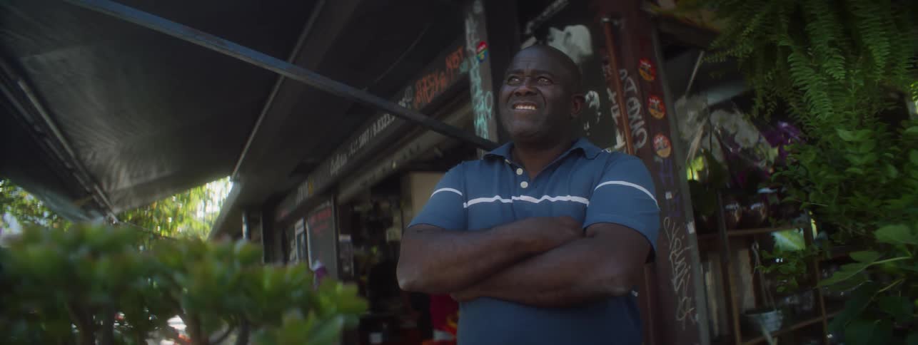 A black man stands with his arms crossed outside a business looking out to the street