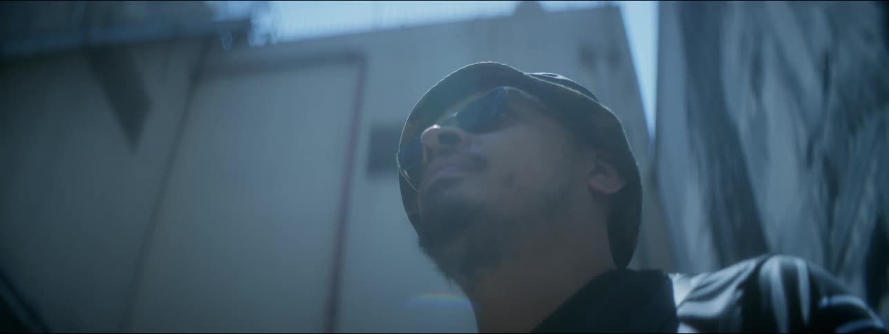 A man in sunglasses and a leather jacket stands in front of a building and looks around before walking off