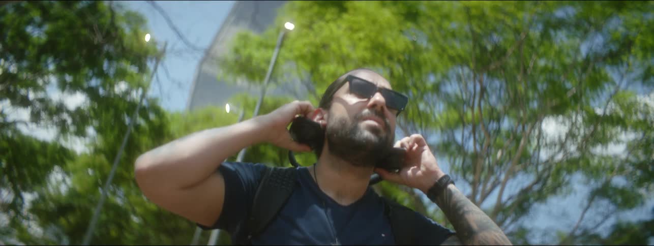 A man wearing sunglasses and headphones is walking on a downtown sidewalk as he takes off the headphones and holds them