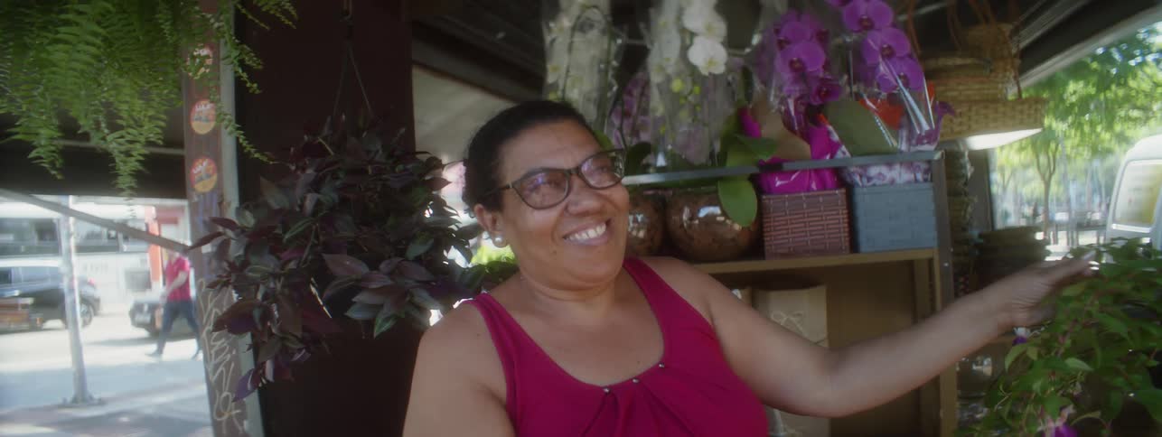 Woman holds plant stem in shop and smiles as male pedestrian walks by in background