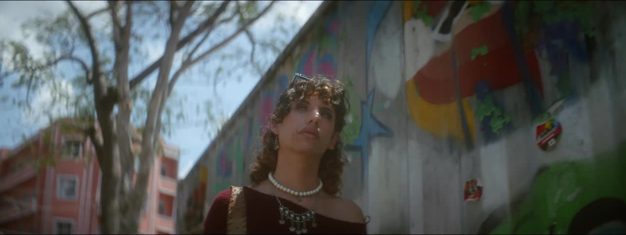 A non-binary teenager walks down a city street by an apartment building and next to an urban art mural