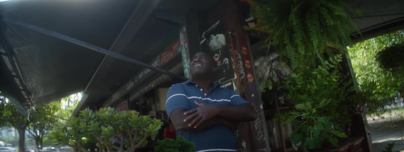 The sun shines brightly as a man stands in front of a business as he folds his arms and looks up