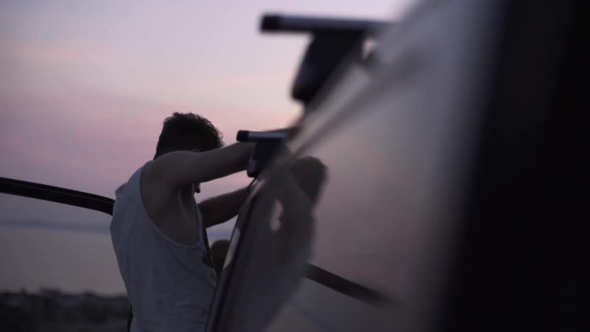 A man adjusting something on the roof of his vehicle as the sun sets at the beach