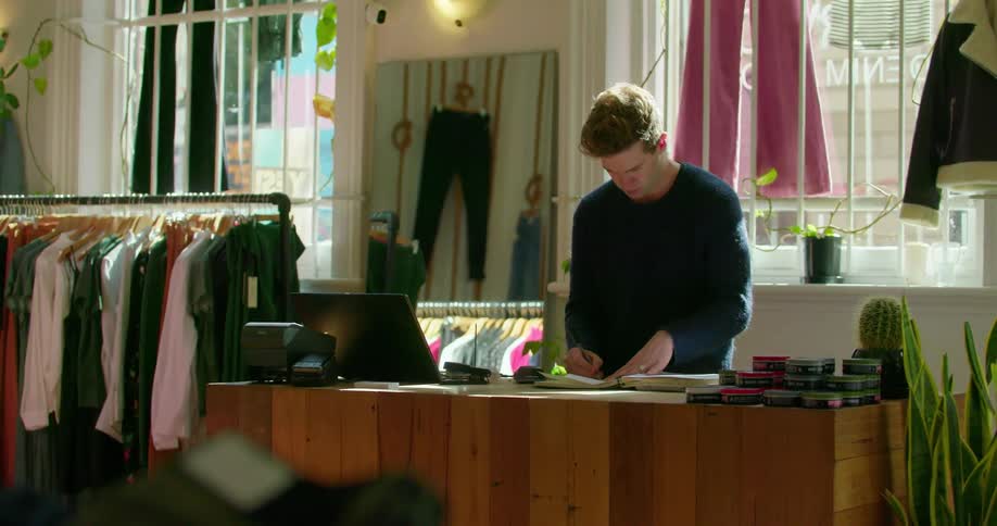 A clothes shop worker writes in a notebook and types on a laptop