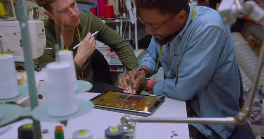 Two men are talking as they look at a tablet while working in a sewing room