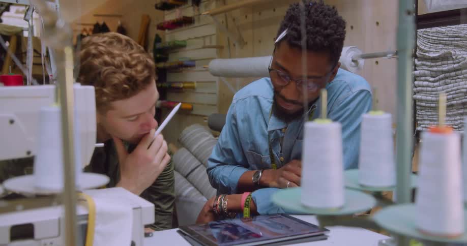 Two men are talking as they look at a tablet while working in a sewing room