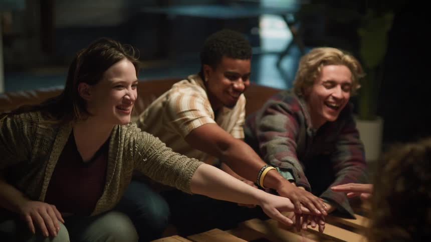 A small group of people are sitting at a table and they all stretch out their arms and put their hands together