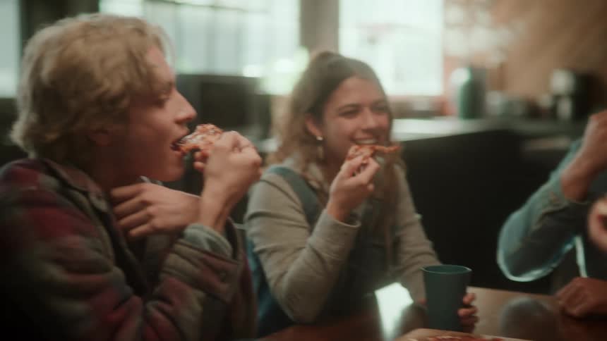 A small group of people are sitting at a table eating pizza in a restaurant and they raise their pizza slices in a toast
