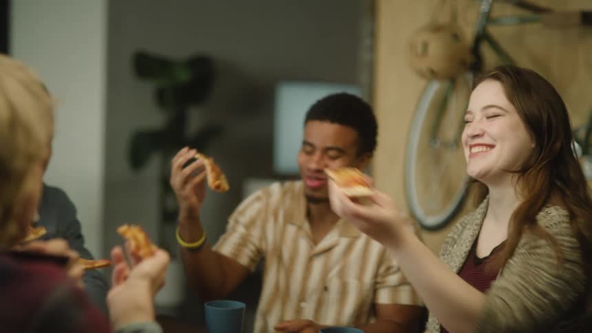 A small group of people are sitting at a table eating pizza in a room with a bicycle on the wall
