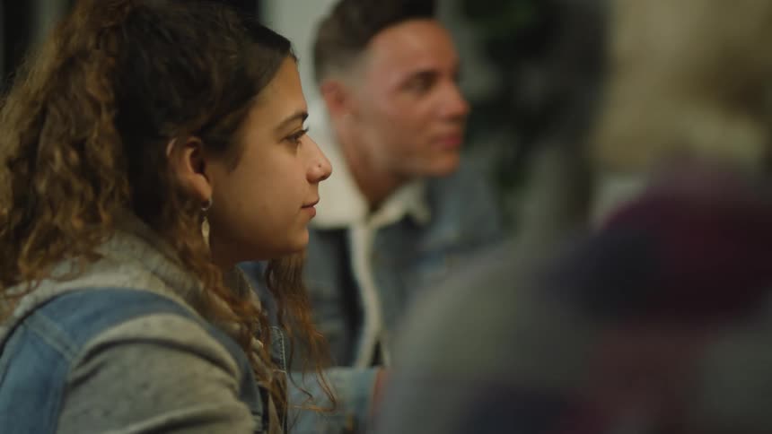 A teenage boy and girl sit at a table in a home and eat while talking with others