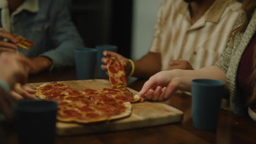 A young man sits down at a table with his friends eating pizza and takes a slice for himself