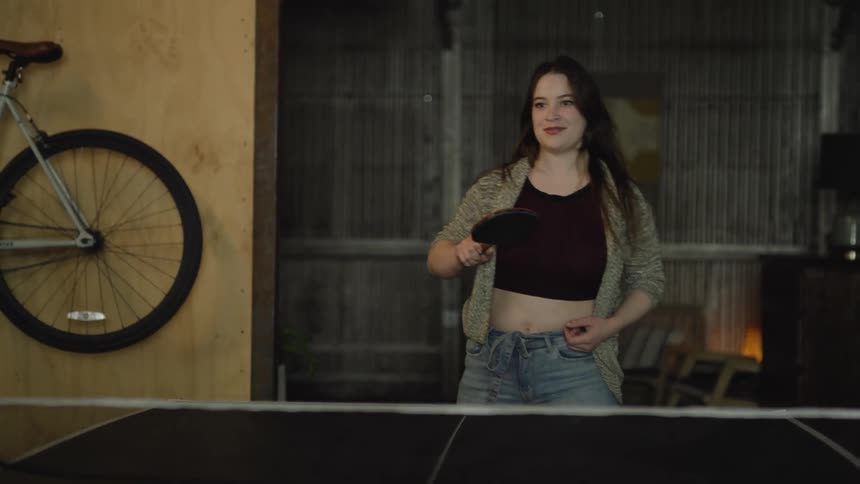 Woman with brown hair black top and jeans plays table tennis in room