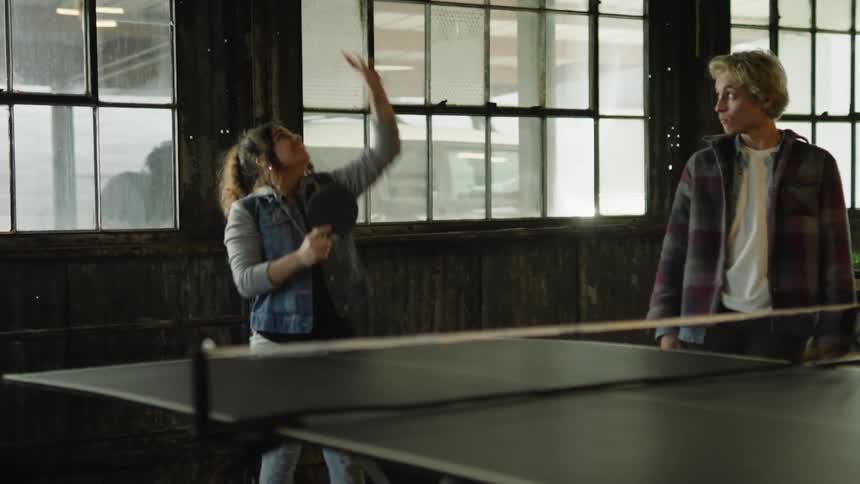 Two teenage boys laugh and catch the ball as they watch a teenage girl play ping pong